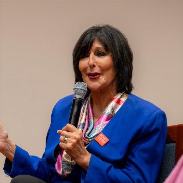 President Mantella speaks into a microphone during a panel discussion. She wears a blue blazer, pink scarf, and a pin that reads �Tech Week�.